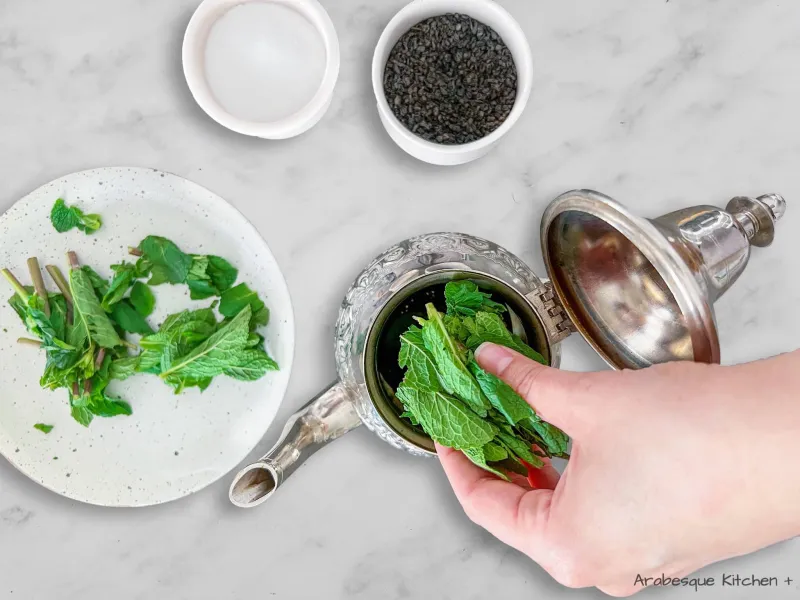 Insert the fresh mint “sprigs”, heaped gunpowder tea and sugar in your Moroccan teapot and fill the teapot with warm water. If the mint sprigs are too large to fit in your pot, cut them in half. 
