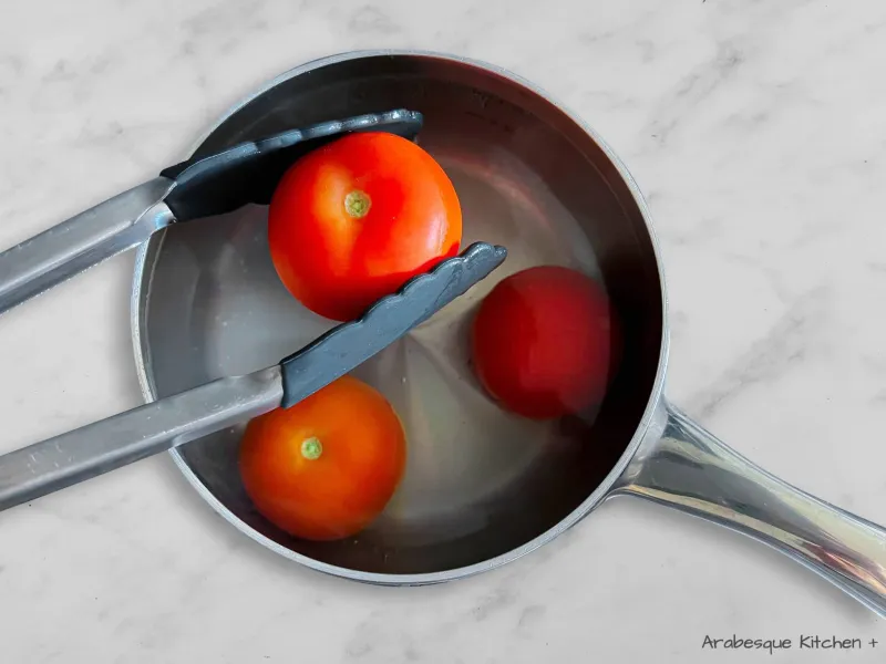 نضع الطماطم في ماء مغلي ونتركها تغلي لمدة 40 ثانية ثم نضعها تحت الماء البارد فهذا سيساعد في تقشير الطماطم. نصفّي الطماطم ونقشرها ونقطعها إلى قطع بسماكة 1 سم.
